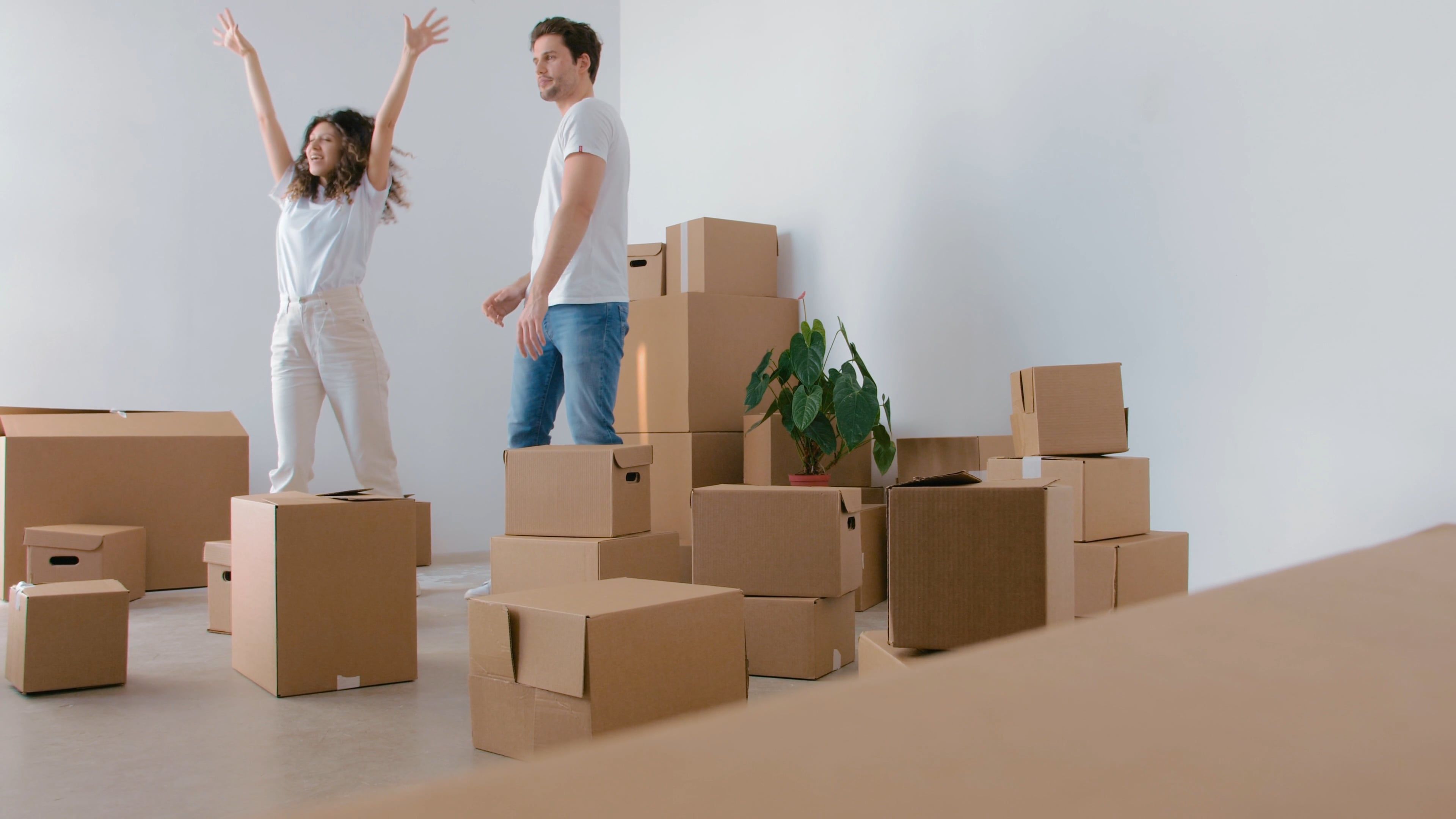 Deux personnes au milieu d’une pièce vide entourée de nombreux cartons de déménagement. Le homme porte un T-shirt blanc et le geste de la femme suggère joie.