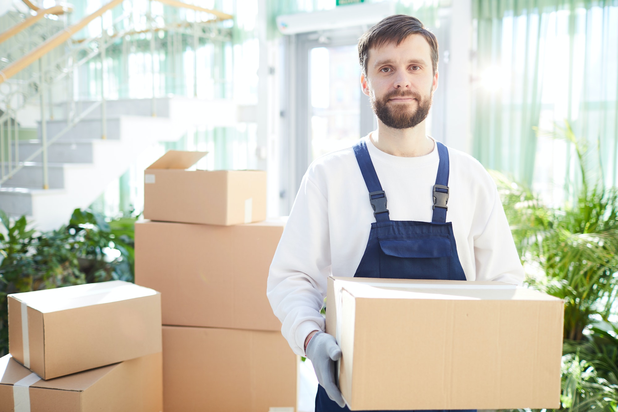 Livreur souriant portant une caisse à l’intérieur, entouré de cartons prêts à déménager, étape déménagement à suivre présentée.
